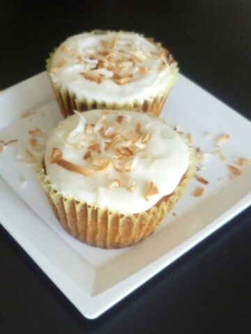 A golden brown cupcake with a white coconut frosting and a slice of pineapple on top. The cupcake is decorated with shredded coconut.