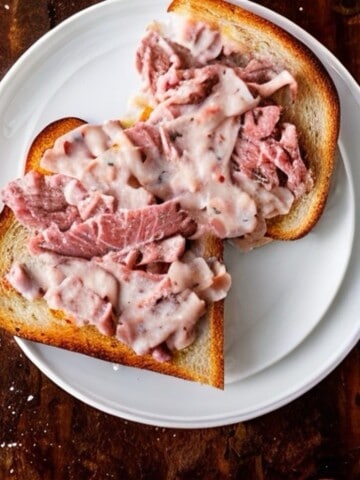 A chipped beef sandwich on white bread with gravy on a white plate on a wooden table