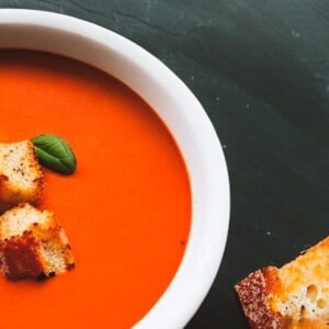 A steaming bowl copycat campbells tomato soup is garnished with a fresh sprig of parsley and accompanied by a grilled cheese sandwich and grilled cheese croutons