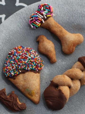 A plate full of homemade gourmet dog treats. The treats are in a variety of shapes, including hearts, ice crem cones, bones, and paw prints. They are decorated with frosting, sprinkles, and carob chips.