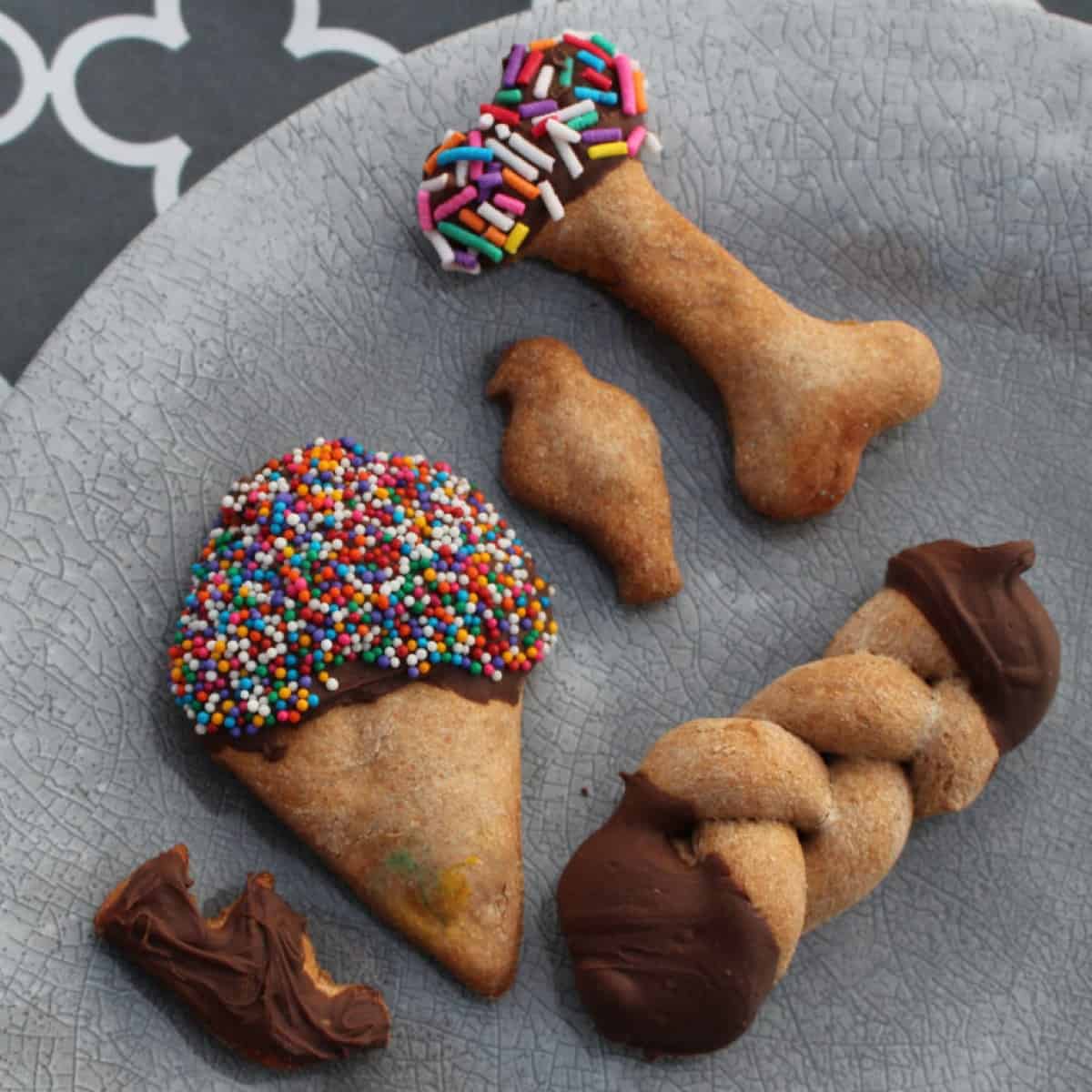 A plate full of homemade gourmet dog treats. The treats are in a variety of shapes, including hearts, ice crem cones, bones, and paw prints. They are decorated with frosting, sprinkles, and carob chips.