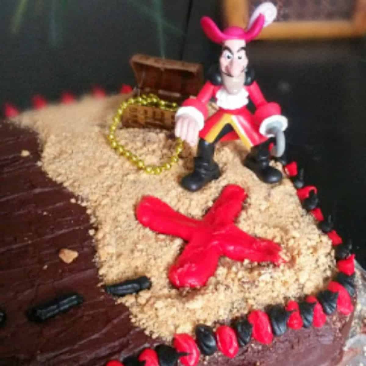 The image shows a chocolate cake with a pirate figurine on top of it. The cake is decorated with a red cross and a treasure chest.