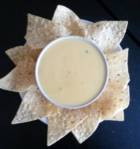 The photo shows a bowl of theater-style nacho cheese sauce without processed cheese. The sauce is smooth and creamy, with a vibrant orange color. It is made from real, high-quality ingredients, resulting in a rich and flavorful taste. The sauce is garnished with finely chopped fresh jalapeños and sprinkled with a dash of paprika, adding a hint of spiciness and an appealing visual appeal. Alongside the bowl of cheese sauce, there are crispy tortilla chips arranged neatly, ready to be dipped into the delicious sauce. The image captures the essence of a mouthwatering snack, perfect for enjoying during a movie night or a casual gathering with friends and family.