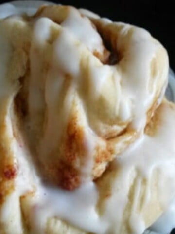 A close up of a cinnamon roll with icing on a plate. The cinnamon roll is fluffy and golden brown, with a generous swirl of cinnamon filling and a drizzle of white icing. The plate is garnished with a few fresh cinnamon sticks.