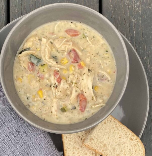 A bowl of Zupas Garden Chowder. The chowder is a light yellow color and contains vegetables such as beans, tomatoes, corn and potatoes. There are also chunks of chicken in the chowder. The chowder is served in a bowl and topped with herbs. 