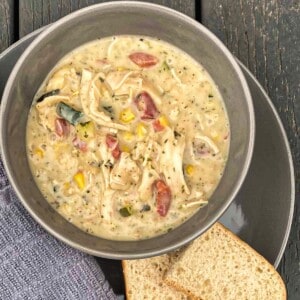 A bowl of creamy chicken chowder topped with shredded cheddar cheese, chopped vegetables like zucchini and corn. The bowl is sitting on a wooden table next to a slice of crusty bread.