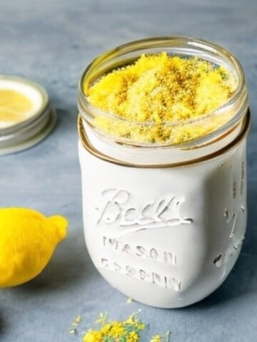A mason jar filled with lemon cake mix from scratch next to a lemon, a lemon slice, and a whisk. The jar is on a white table with a wooden background.