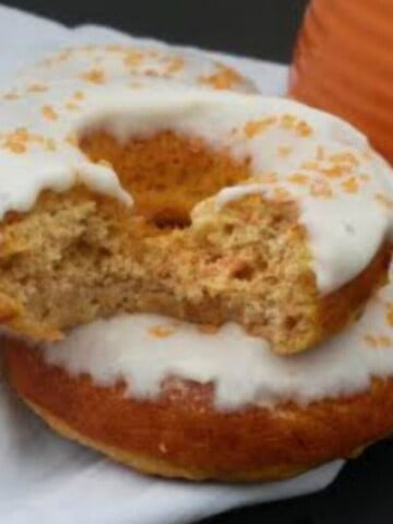 Two carrot cake donuts stacked on top of each other, with a bite taken out of one of them. The donuts are frosted with a cream cheese frosting