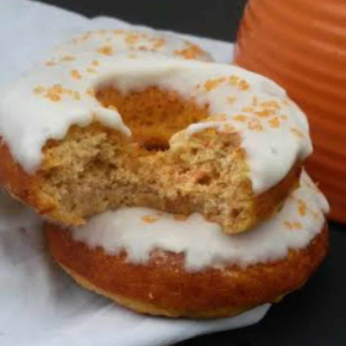 Two carrot cake donuts stacked on top of each other, with a bite taken out of one of them. The donuts are frosted with a cream cheese frosting 