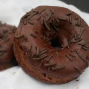 A chocolate baked donut with a chocolate glaze and chocolate sprinkles. The donut is sitting on a white plate.