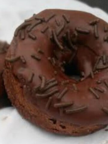 a picture of a chocolate donut with chocolate frosting. Chocolate sprinkles are on top of the donut. The donut is brown with a white background.