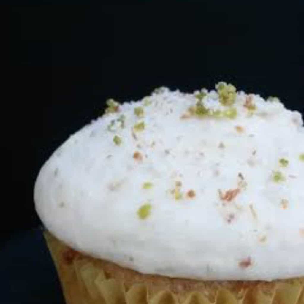a picture of a pineapple cupcake with toasted coconut and lime zest on top. The cupcake is white and the background is black. 