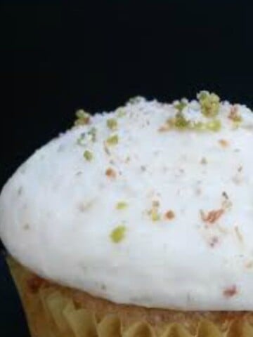 a picture of a pineapple cupcake with toasted coconut and lime zest on top. The cupcake is white and the background is black.