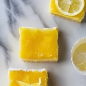 A close-up photo of a plate of lemon curd bars. The bars have a buttery shortbread crust and a bright yellow lemon curd filling. The top of the bars is dusted with powdered sugar and garnished with a lemon slice.