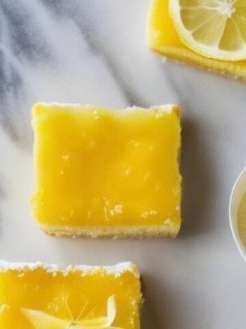 A close-up photo of a plate of lemon curd bars. The bars have a buttery shortbread crust and a bright yellow lemon curd filling. The top of the bars is dusted with powdered sugar and garnished with a lemon slice.