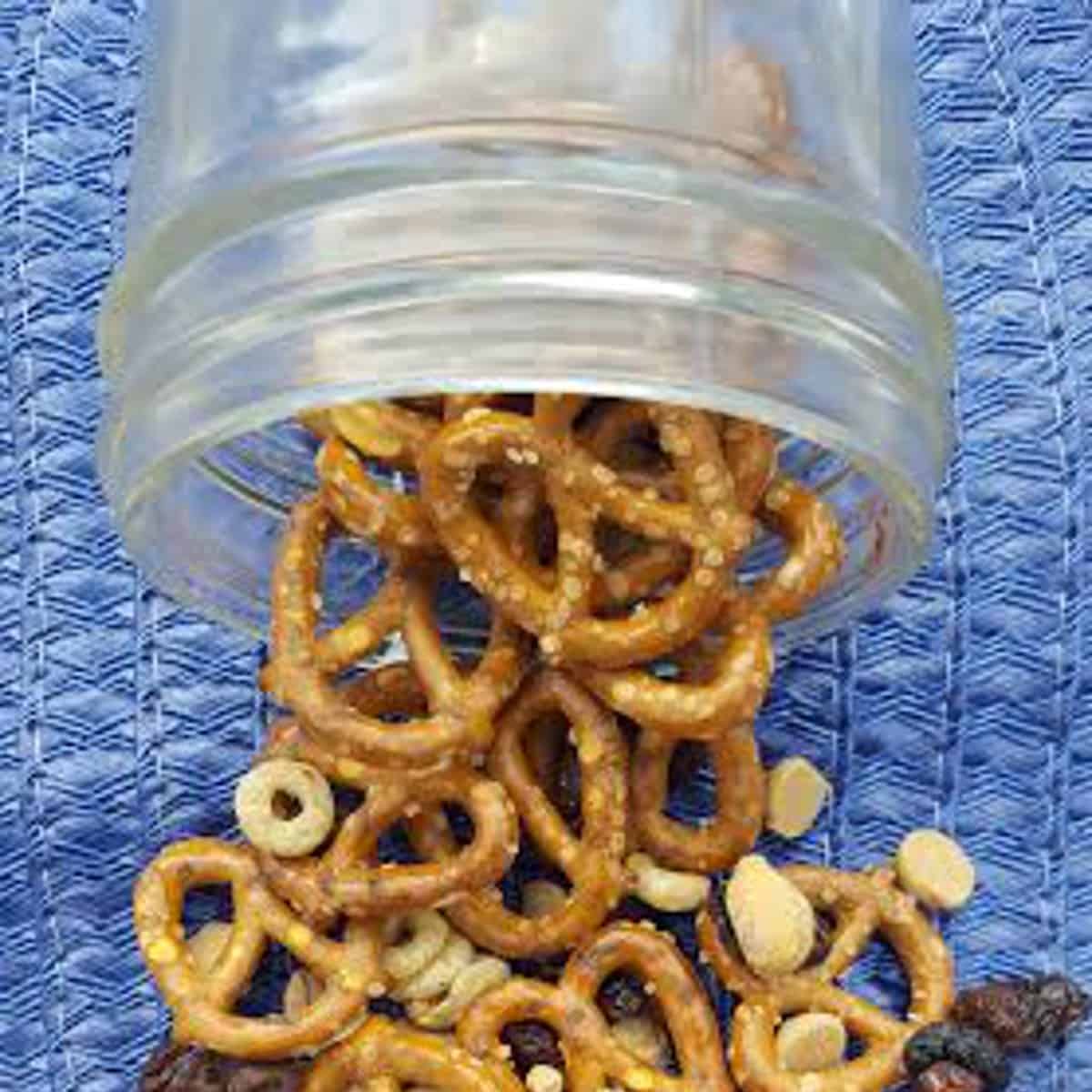 A close up photo of trail mix with chocolate spilling out a mason jar. The jar contains pretzels, cheerios, nuts, dried fruit and chocolate chips.