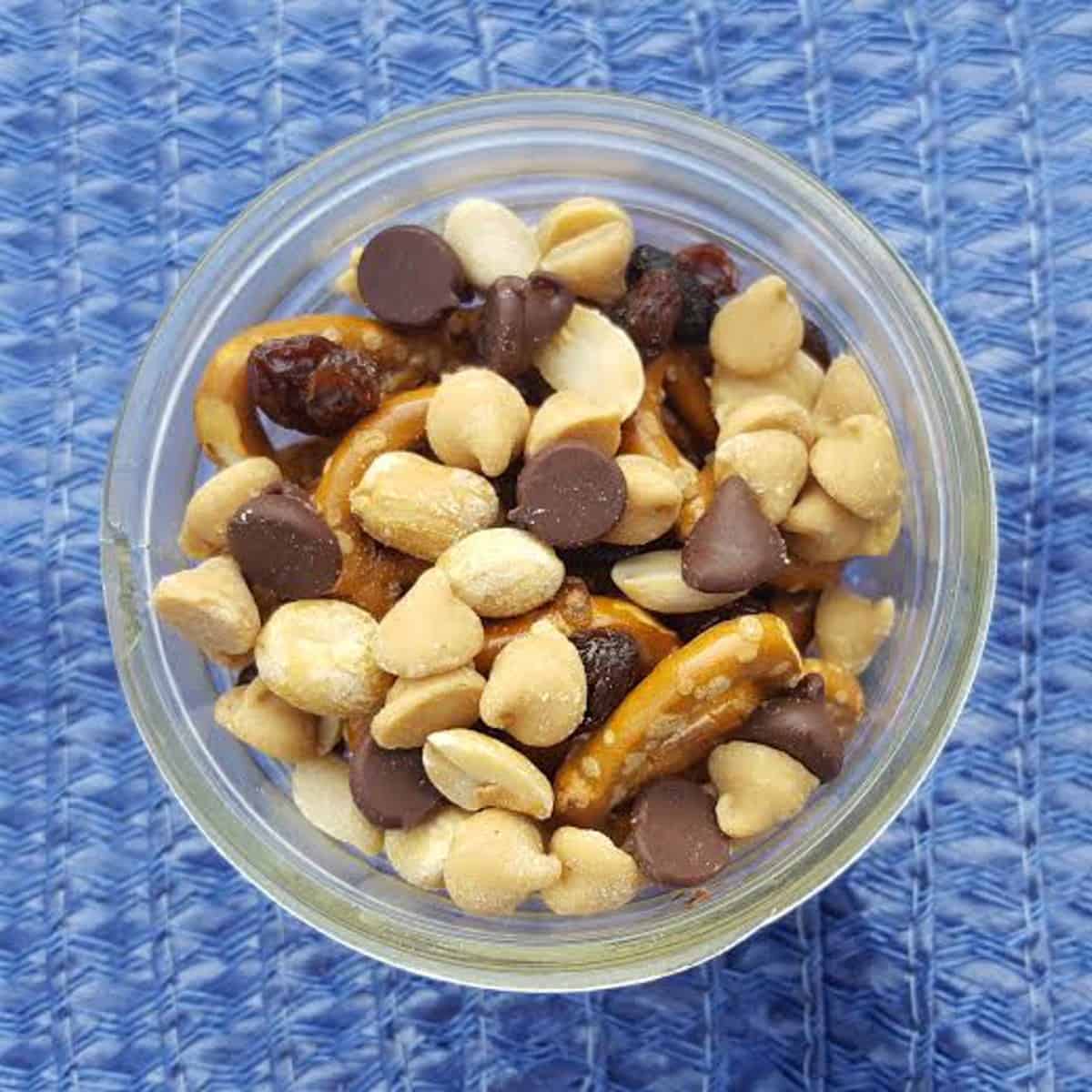 Dark Chocolate trail mix in a mason jar on a blue rustick background. The trail mix contains nuts, fruits, and seeds. It also contains dark chocolate chips and pretzels