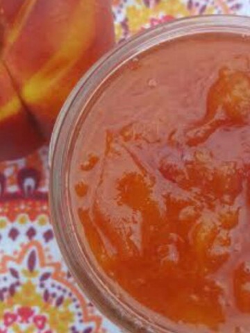A close-up photo of a jar of nectarine jam on a table. The jam is a deep orange color and has a slightly glossy sheen. The jar is labeled "Nectarine Jam" and has a lid.