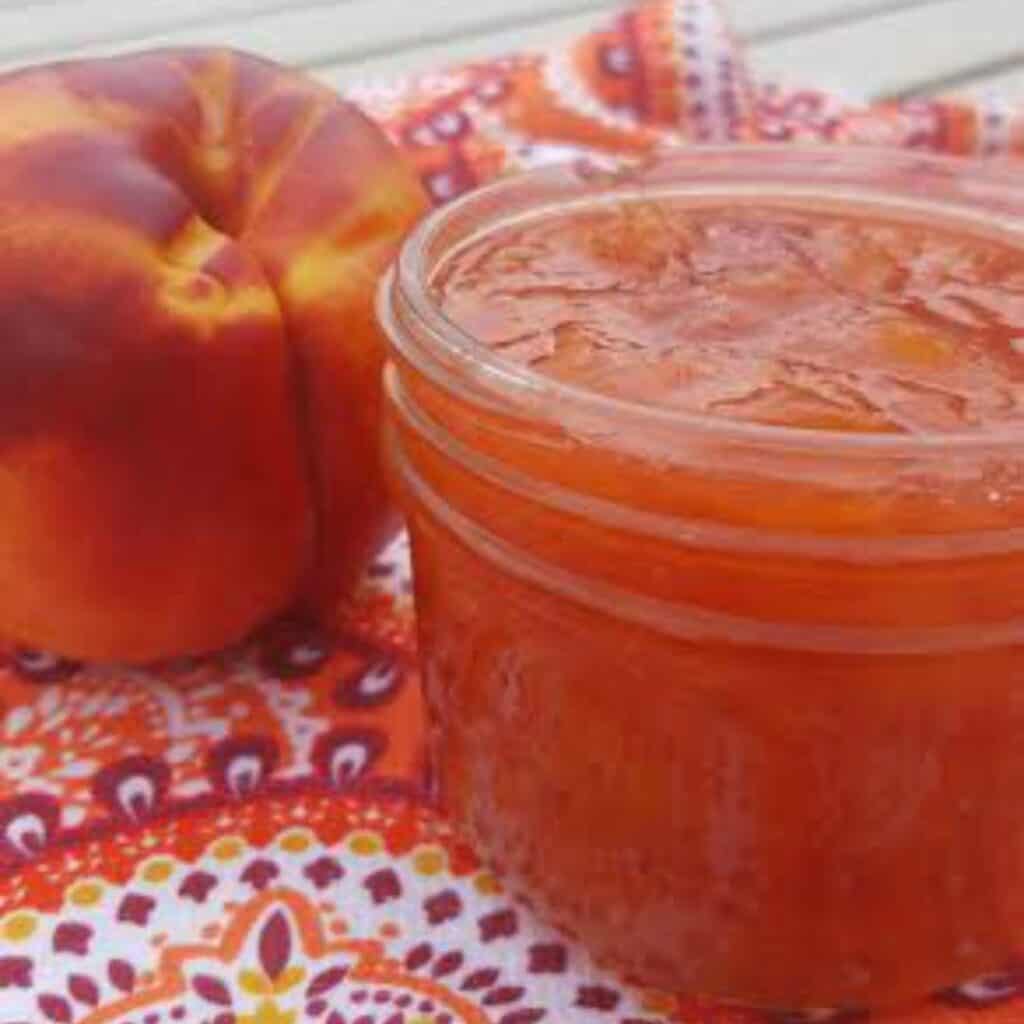 a jar of orange nectarine jam recipe. A nectarine is pictured in the back of the jar of jam. 