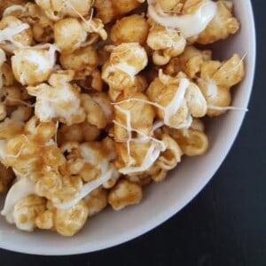 A close-up photo of a bowl of apple caramel popcorn. The popcorn is coated in a thin layer of caramel sauce and has a drizzle of melted white chocolate