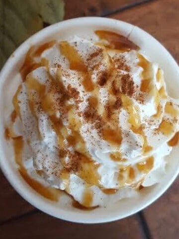 A close-up photo of a glass of caramel apple cider with a cinnamon stick. The cider is a deep amber color with a thick and creamy consistency. There is a swirl of caramel sauce on the side of the glass and a sprinkle of cinnamon and a dollop of whipped cream on top.