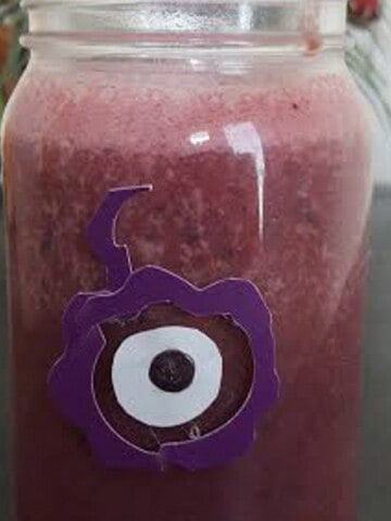 A close-up photo of a glass filled with a purple smoothie. The smoothie has a thick and creamy consistency, and is topped with a sprinkle of chia seeds and a few blueberries. The glass is sitting on a white table, and there is a straw in the smoothie.