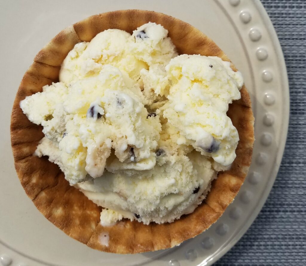 a delicious cookie dough ice cream recipe shows a bowl of vanilla ice cream filled with chocolate chip cookie dough chunks in a waffle bowl 