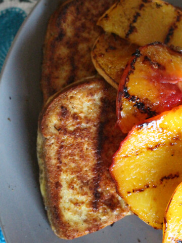 Golden-brown grilled French toast topped with grilled peaches and served with a side of maple syrup. The bread is perfectly toasted with crisp edges and a soft, fluffy interior