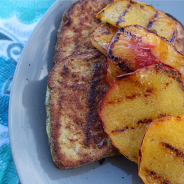 Grilled French toast topped with caramelized peaches, and a drizzle of maple syrup. The peaches are beautifully golden and juicy, complementing the golden-brown grilled bread slices.