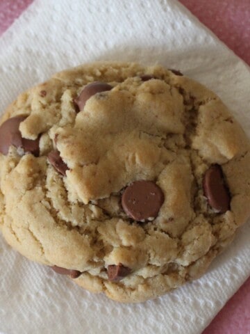 A close-up view reveals a giant, golden brown Crumbl chocolate chip cookie resting on a parchment-lined baking sheet.
