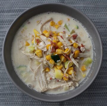 This image shows a bowl filled with a steaming bowl of chicken corn chowder. The chowder is a creamy and rich soup with a golden-yellow color. It is topped with a garnish of chopped fresh herbs, which adds a pop of green to the dish. The soup is brimming with ingredients such as tender pieces of chicken, sweet corn kernels, diced potatoes, carrots, and onions, all immersed in a thick and velvety broth. The steam rising from the bowl indicates its warmth, making it an inviting and comforting meal option. The image captures the essence of a hearty and delicious chicken corn chowder, perfect for a satisfying meal.
