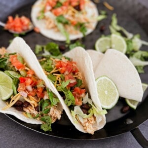 A close-up photo of three soft corn tortillas piled high with shredded chicken, black beans, diced tomatoes, shredded cheese, and a dollop of sour cream. Cilantro leaves add a pop of freshness, and the crock pot used to make these crock pot chicken ranch tacos sits in the background