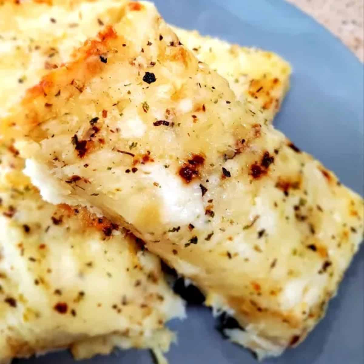 A close-up photo of a rectangular piece of golden brown Italian cheese bread on a blue plate. The bread is cut into squares, revealing melted mozzarella cheese and flecks of herbs in the center. A light dusting of parmesan cheese adorns the top
