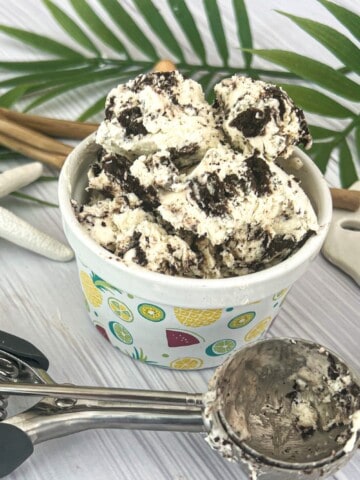 A bowl of heavy whipping cream ice cream with chocolate cookie crumbles mixed in, next to a silver ice cream scooper. The ice cream is in a white ceramic bowl and sits on a light-colored wooden table.