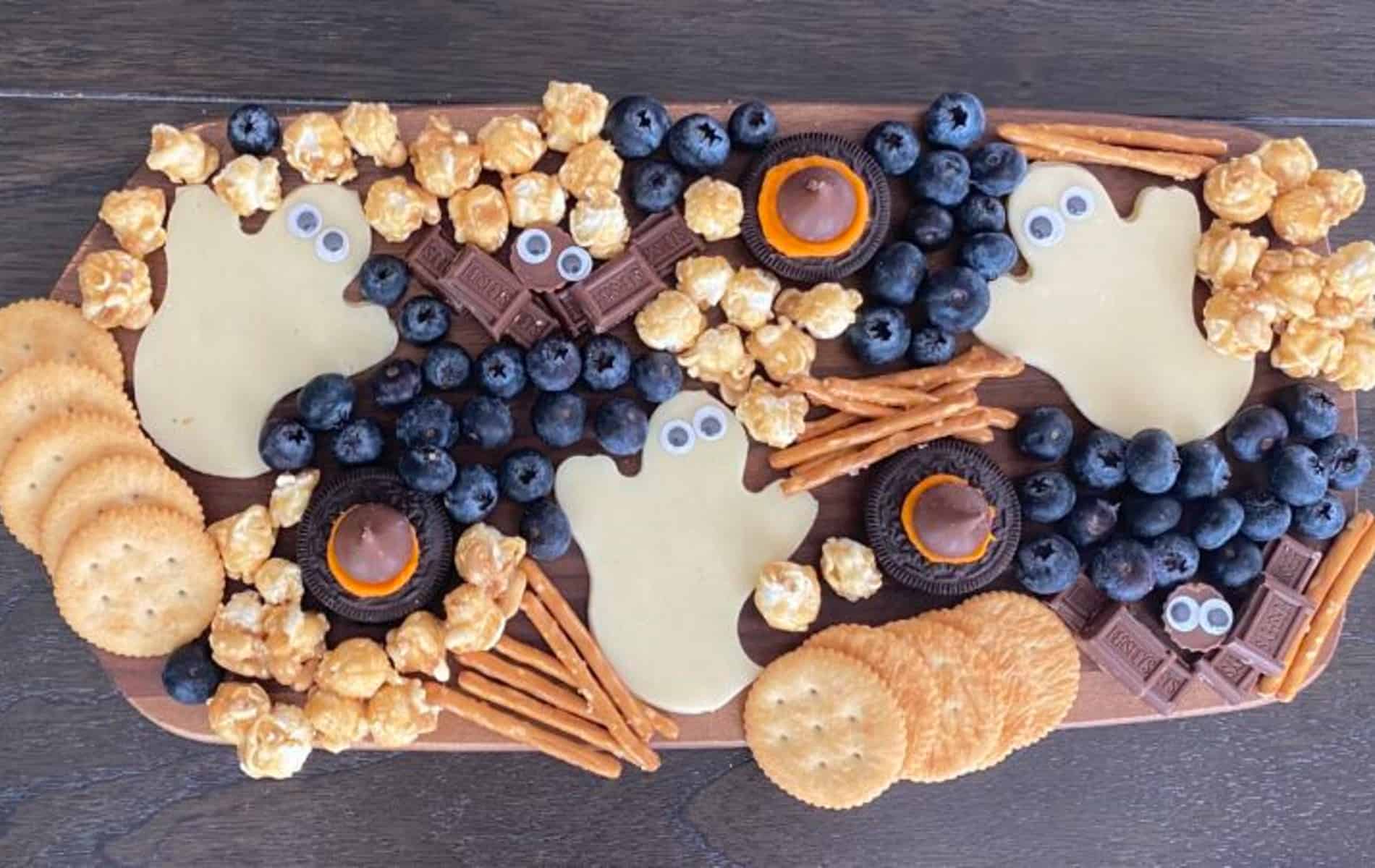 an aerial shot of a Halloween snack board with cheese slices blueberries, cookies, crackers, popcorn and pretzels. 