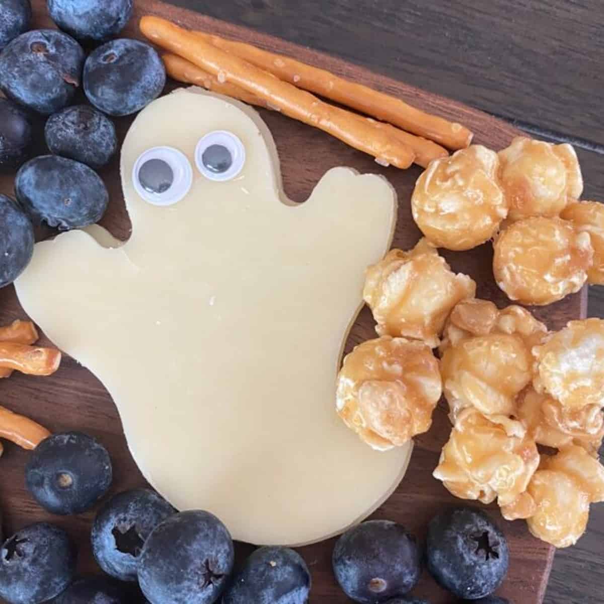A slice of cheese cut to resemble a ghost. A pair of googly eyes are added to the cheese ghost. The cheese ghost is arranged on a Halloween snack board. 