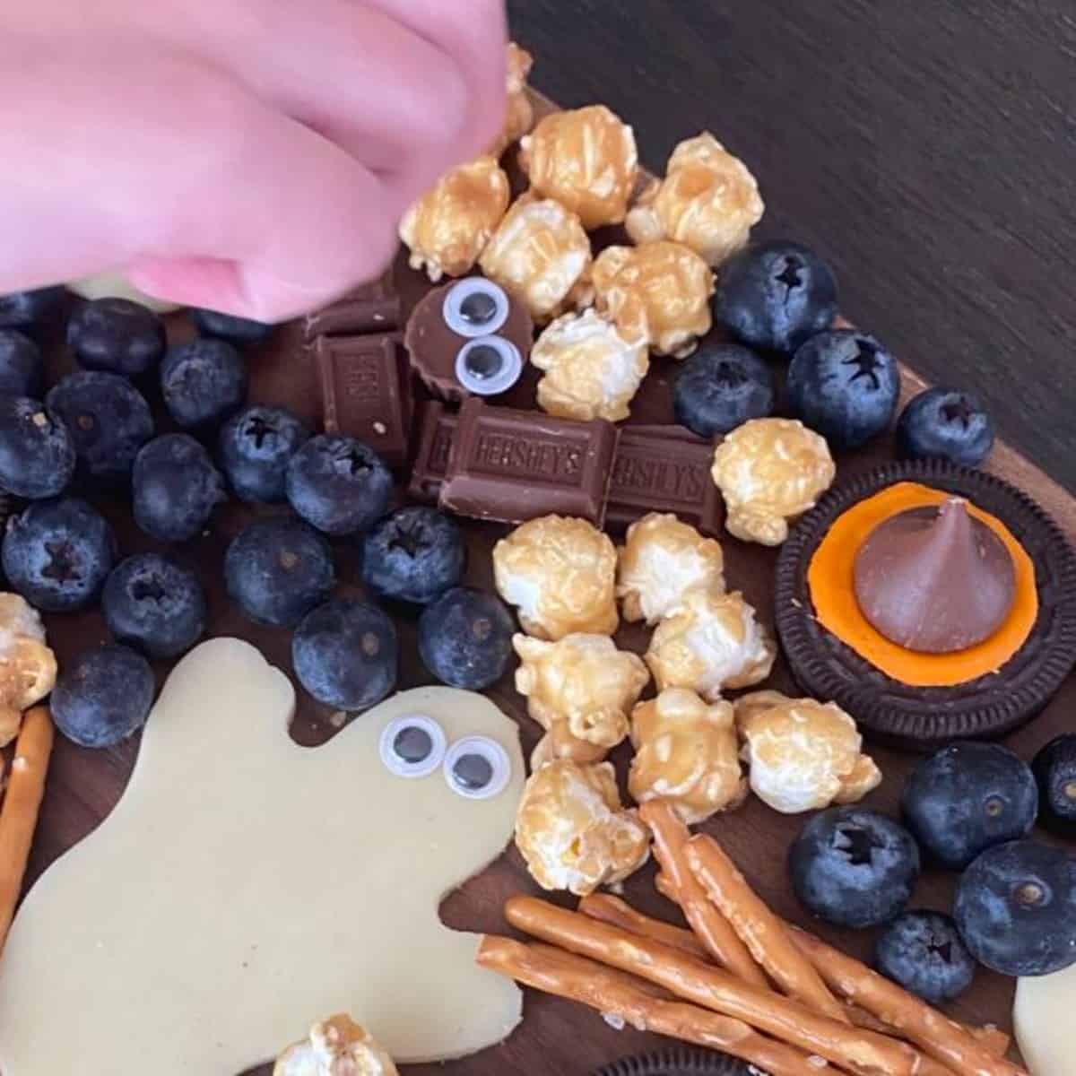 A Halloween dessert board with witches hat cookies with Oreos, cut out cheese ghosts and chocolate bats. The board also has caramel popcorn, blueberries and pretzels.