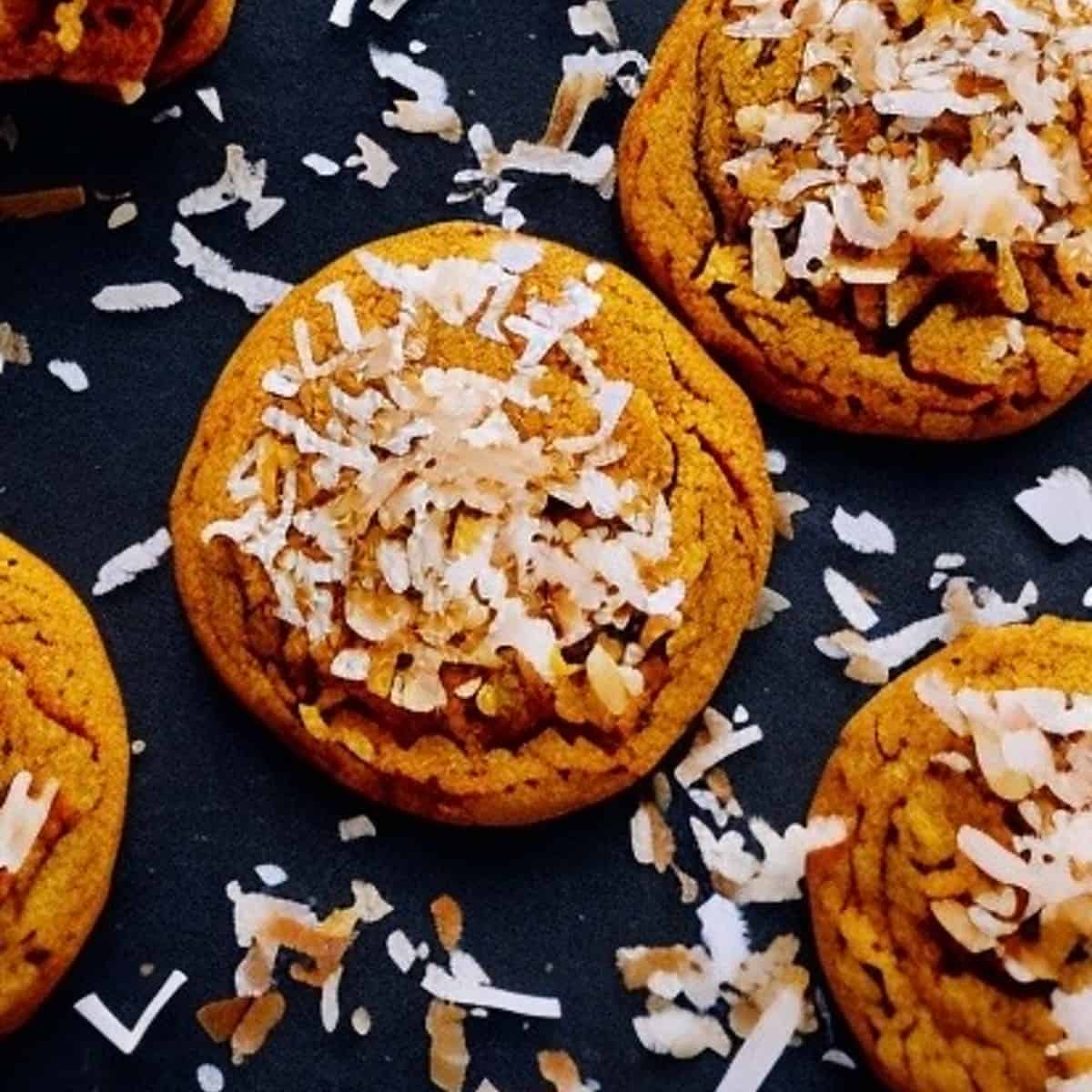 A close-up photo of several pumpkin coconut cookies on a white plate. The cookies are round and golden brown, with cracks on the surface. They are sprinkled with shredded coconut