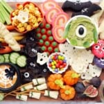 A close-up photo of a Halloween charcuterie board filled with sweet and savory treats arranged in sections on a black serving platter. Spooky elements like cobwebs and spiders add to the festive atmosphere.