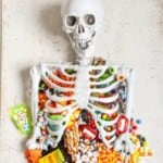A festive Halloween display featuring a human skeleton playfully positioned on a table, its ribcage filled with an assortment of colorful candy. The candy includes lollipops, chocolate bars, gummy bears, and candy corn, creating a visually appealing and sugary spectacle. The skeleton's arms rest playfully on the table's edge, further adding to the lighthearted and celebratory atmosphere.