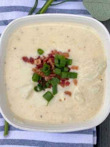 A close-up photo of a bowl of Chili's Baked Potato Soup. The soup is creamy and white, with visible chunks of potato, bacon, and green onions. The bowl is garnished with a dollop of sour cream, shredded cheddar cheese, and a sprinkle of paprika. The soup is served in a red ceramic bowl with a white rim, and there is a metal spoon resting on the edge of the bowl.
