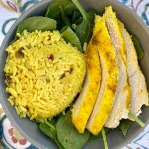 A photo of a dish containing saffron chicken and rice with spinach. The chicken pieces are golden brown and appear to be bone-in, skin-on thighs. They are situated on a bed of fluffy yellow rice that has been seasoned with saffron, giving it a vibrant color. There are also green spinach leaves scattered around the dish, adding a pop of color and freshness.