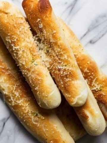 A close-up photo of several gluten-free breadsticks on a white plate. The breadsticks are long and thin, with a golden brown color and a slightly irregular, rustic appearance. They are arranged vertically on the plate, and some are slightly bent or leaning against each other. There are a few small crumbs scattered on the plate around the breadsticks.