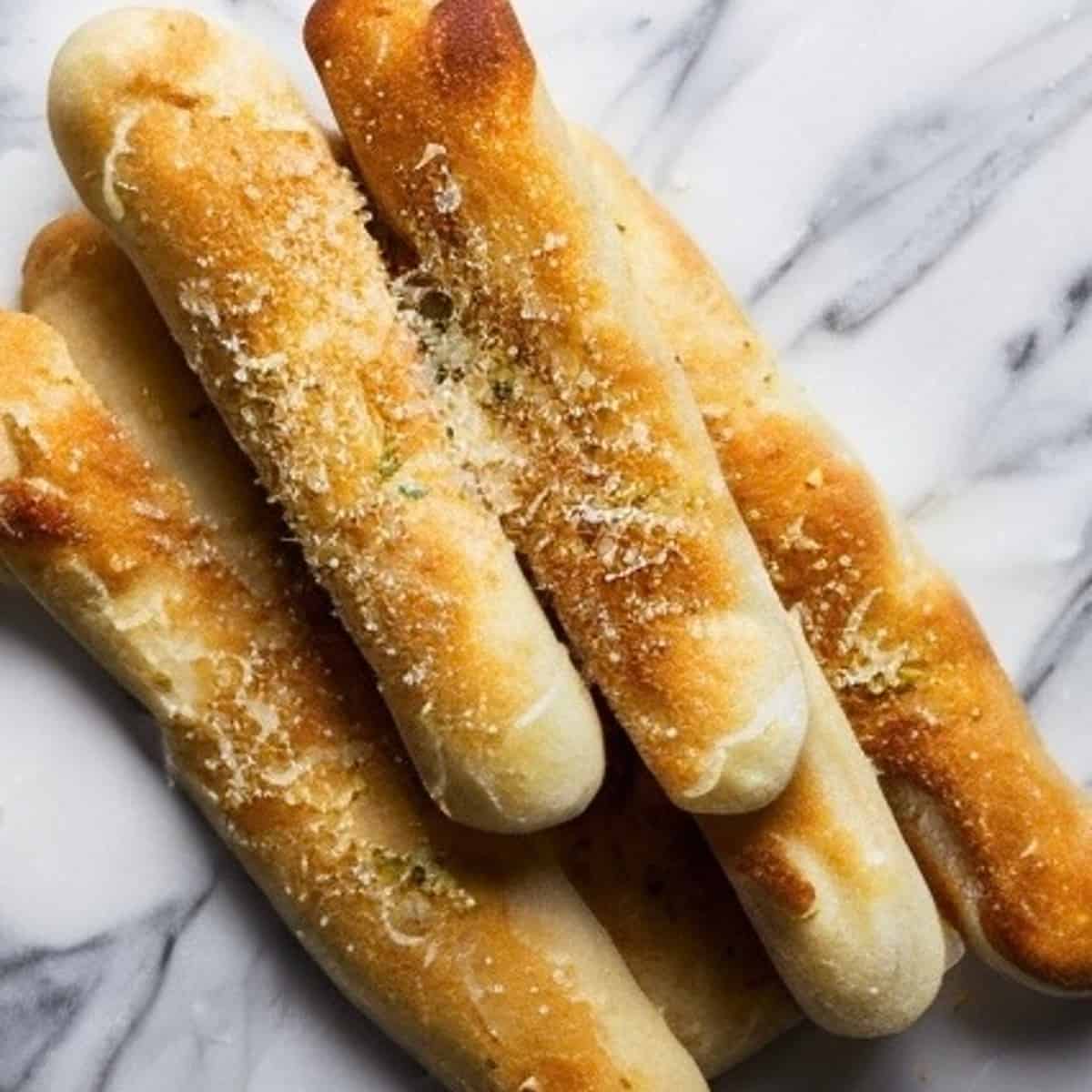 A close-up photo of several gluten-free breadsticks on a white plate. The breadsticks are long and thin, with a golden brown color and a slightly irregular, rustic appearance. They are arranged vertically on the plate, and some are slightly bent or leaning against each other. There are a few small crumbs scattered on the plate around the breadsticks.