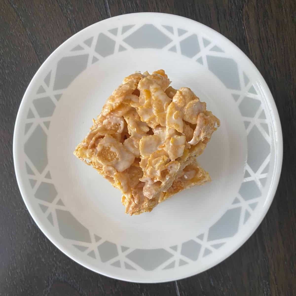 A close-up photo of a plate filled with homemade Frosted Flake treats. The treats are square-shaped and golden brown, with visible pieces of frosted corn flakes. They are arranged in a single layer on a white plate with a patterned blue rim.