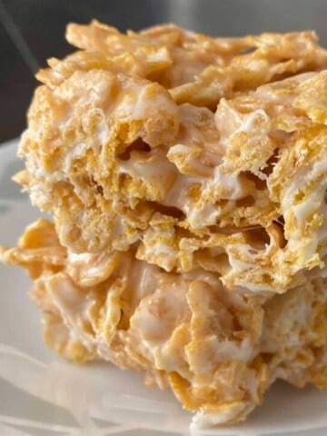 A close-up photo of a plate filled with Frosted Flakes Rice Krispie Treats. The treats are square-shaped and golden brown, with visible pieces of Frosted Flakes cereal throughout. They are arranged in a single layer on the plate.