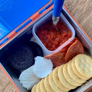 A lunchbox filled with a variety of homemade pizza lunchables ingredients for assembling mini pizzas. The lunchbox is open, revealing compartments containing pizza sauce, cookies, cheese rounds, pepperoni and butter crackers.