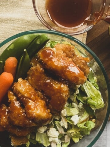 A close-up of a bowl of Wingers sticky fingers recipe with a side of shredded carrots and lettuce on a wooden table. The wings are coated in a red sauce and appear saucy and glistening.