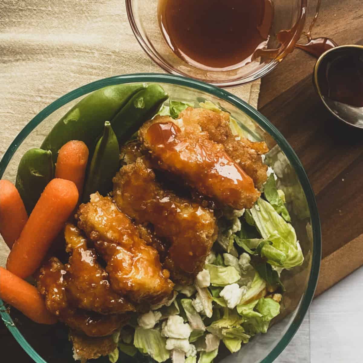 A close-up of a bowl of Wingers sticky fingers recipe with a side of shredded carrots and lettuce on a wooden table. The wings are coated in a red sauce and appear saucy and glistening.