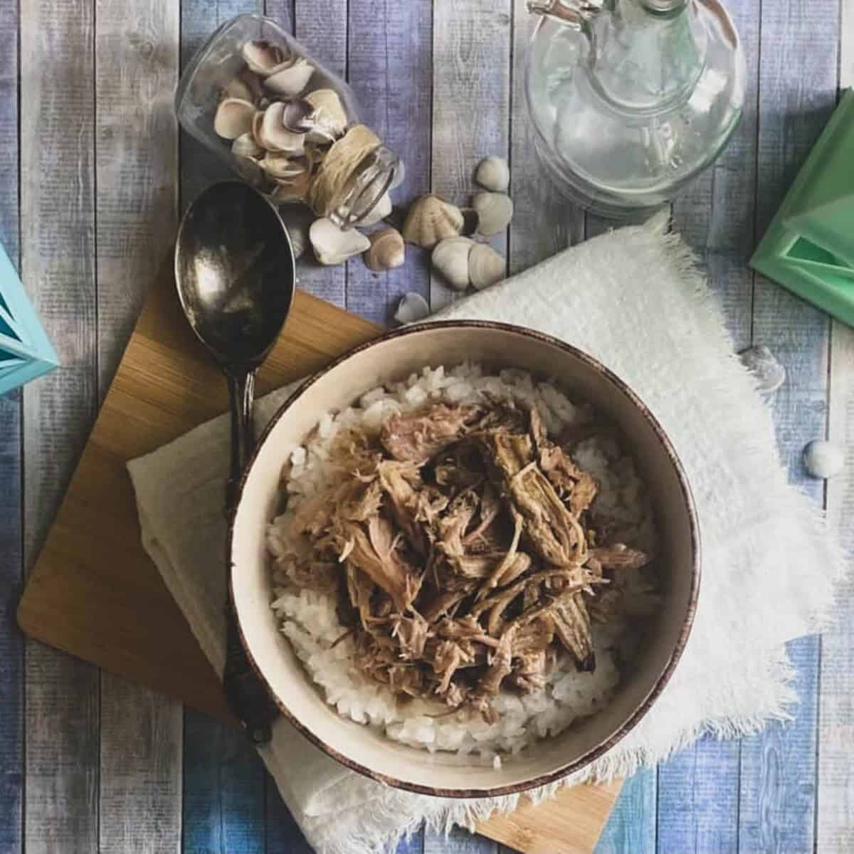 A close-up of a wooden table with a bowl of shredded brown pulled smoked kalua pork on top of white rice.